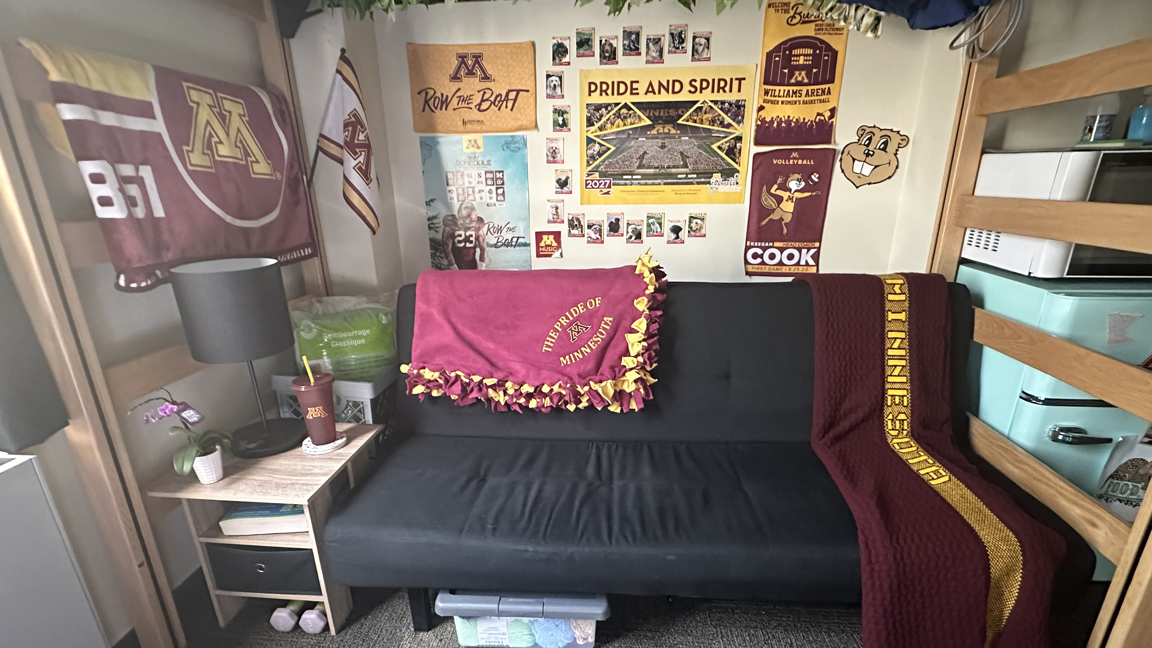 2024 Goldy Room. A black futon under a lofted bed with two maroon U of M blankets on it. U of M posters on the wall behind.