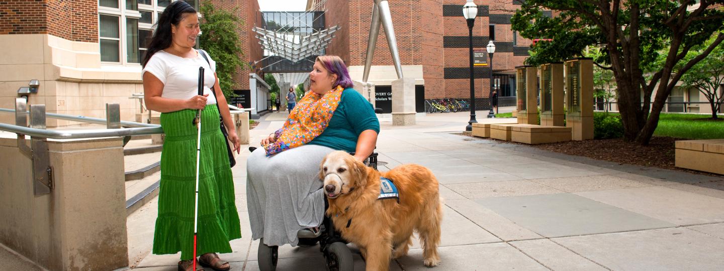 A student with a white cane talks to a student using a wheelchair and service dog.
