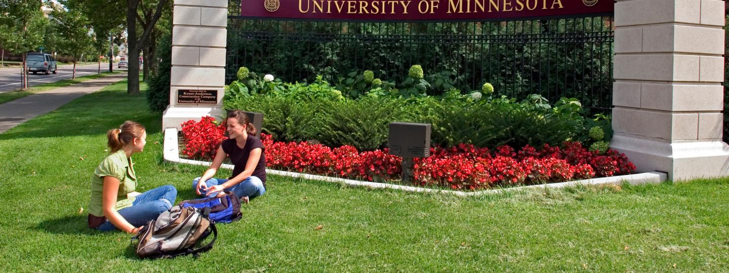Two students sit in the grass at a U of M gateway marker near Wilkins Hall