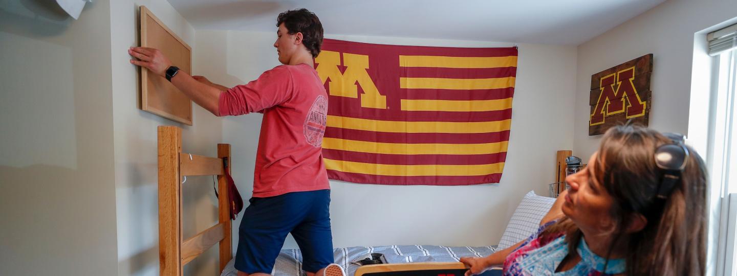 A student is kneeling on their bed while hanging a bulletin board on their wall while another person leans away to examine if it is straight
