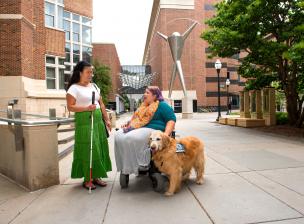 A student with a white cane talks to a student using a wheelchair and service dog.