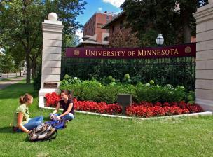 Two students sit in the grass at a U of M gateway marker near Wilkins Hall