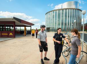 UMPD police members, one in a grey polo and black bike helmet and another in officer's uniform, chat with a student at a bike rack near the Washington avenue bridge