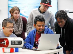 Several people in a group are looking at a laptop screen over a person's shoulder
